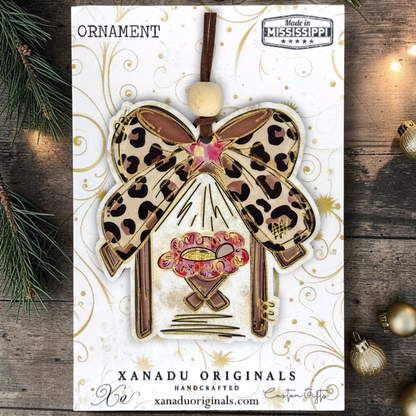 A handmade Christmas ornament by Xanadu Originals, featuring a leopard print bow, a pink and gold floral center, and a brown hanging cord, presented on its white printed backing card with holiday-themed flat-lay props.