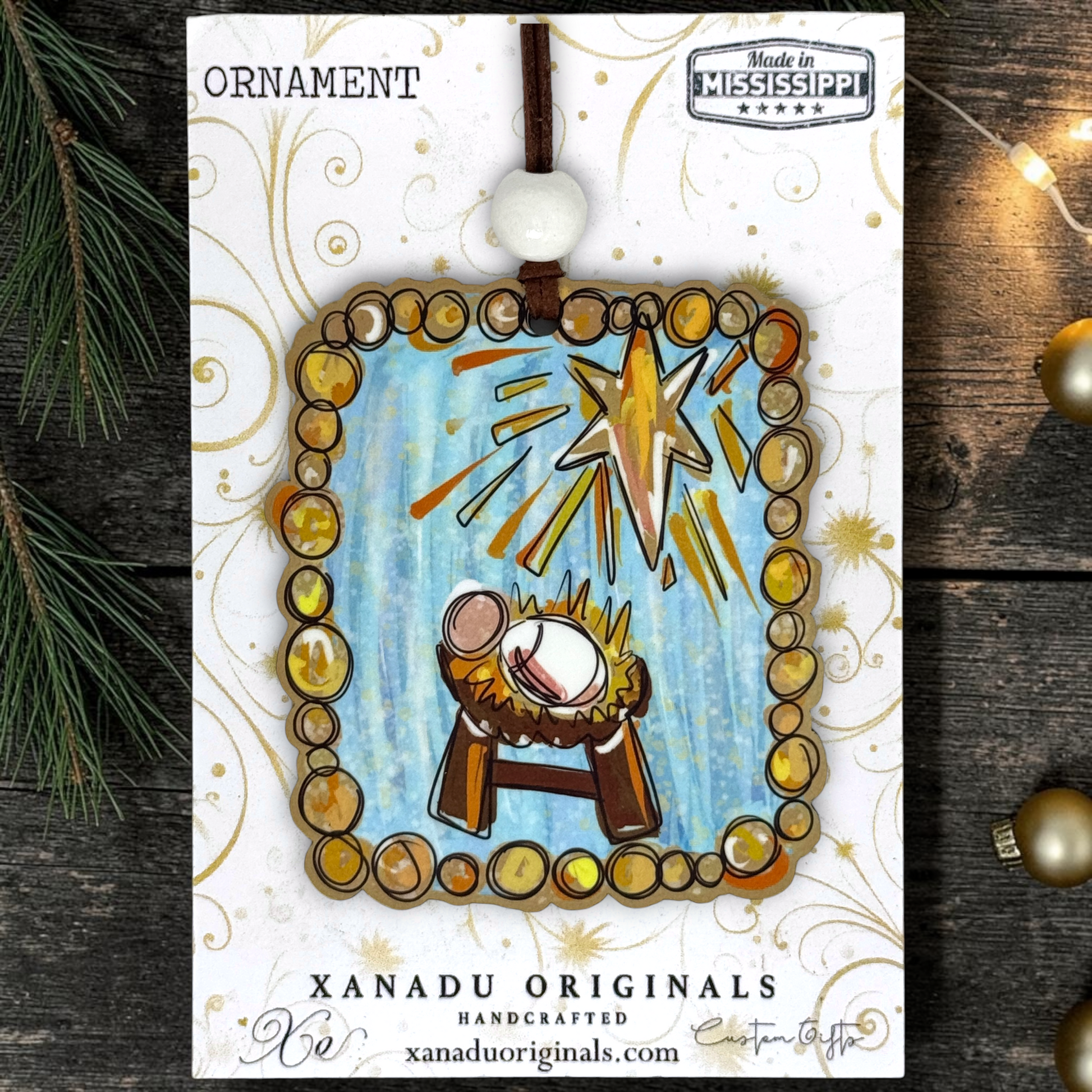 A handmade Christmas ornament card from Xanadu Originals, featuring a nativity scene ornament with a gold star, baby Jesus in a manger, and a circular border made of gold and brown beads. The card is displayed on a dark wooden surface with pine needles and holiday lights.