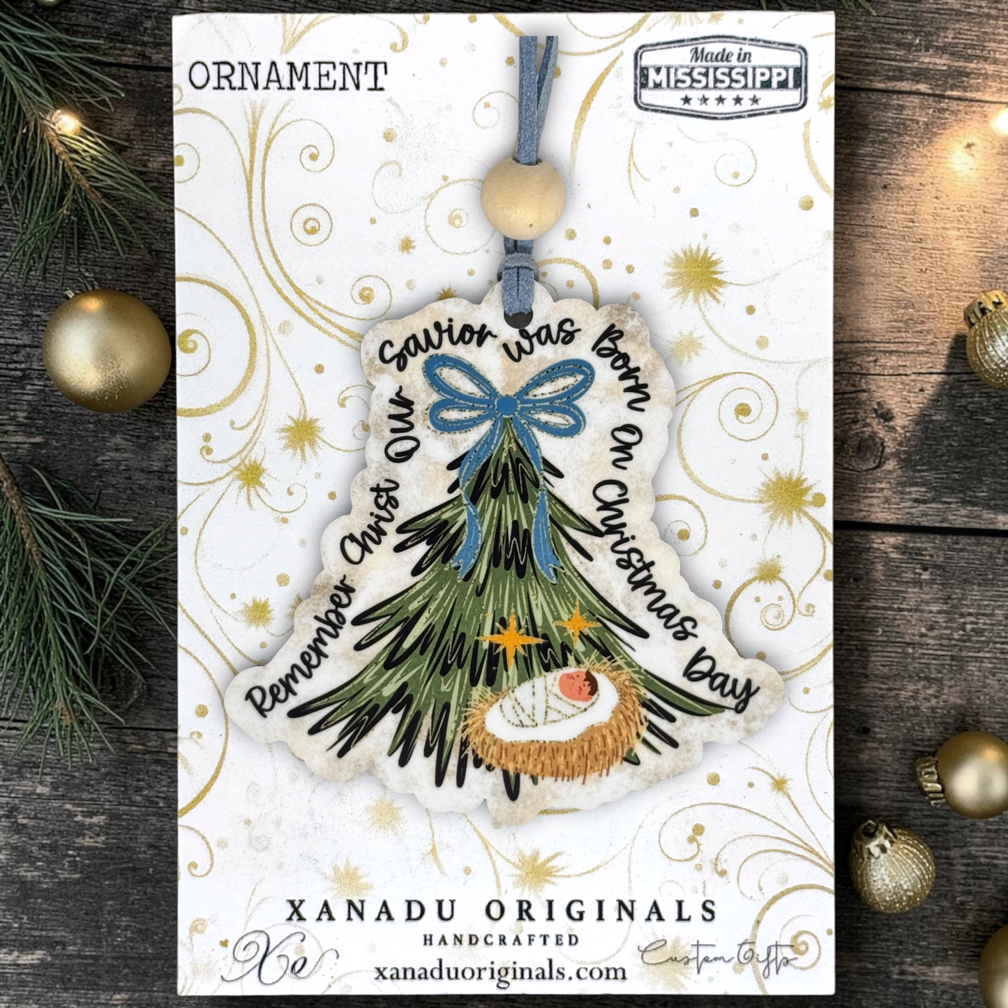 Handcrafted Christmas ornament card featuring a nativity tree ornament with a blue bow, gold star accents, and the text 'Savior was born on Christmas Day' and 'Remember Christ Our Savior was born on Christmas Day.' The card is displayed on a rustic dark wood background with pine branches and gold ornaments.