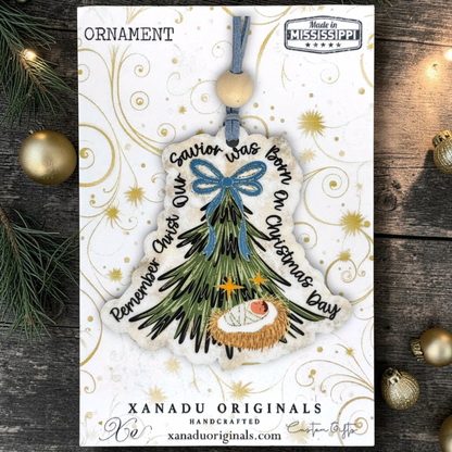 Handcrafted Christmas ornament card featuring a nativity tree ornament with a blue bow, gold star accents, and the text 'Savior was born on Christmas Day' and 'Remember Christ Our Savior was born on Christmas Day.' The card is displayed on a rustic dark wood background with pine branches and gold ornaments.