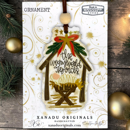 A handmade Christmas ornament shaped like a stable, featuring the text 'the weary world rejoices' in black script with gold and red accents, displayed on its white branded backing card over a rustic wood background with pine branches and gold ornaments.