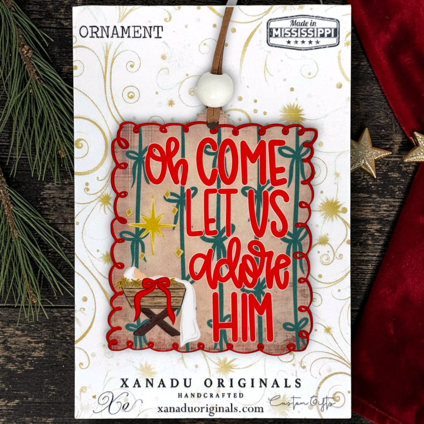 A Christmas ornament carded on a branded display card. The ornament is a square wooden plaque with red scalloped trim, red lettering reading “OH COME LET US adore HIM,” green vertical bars, gold starburst accents, and a small baby Jesus near the bottom.