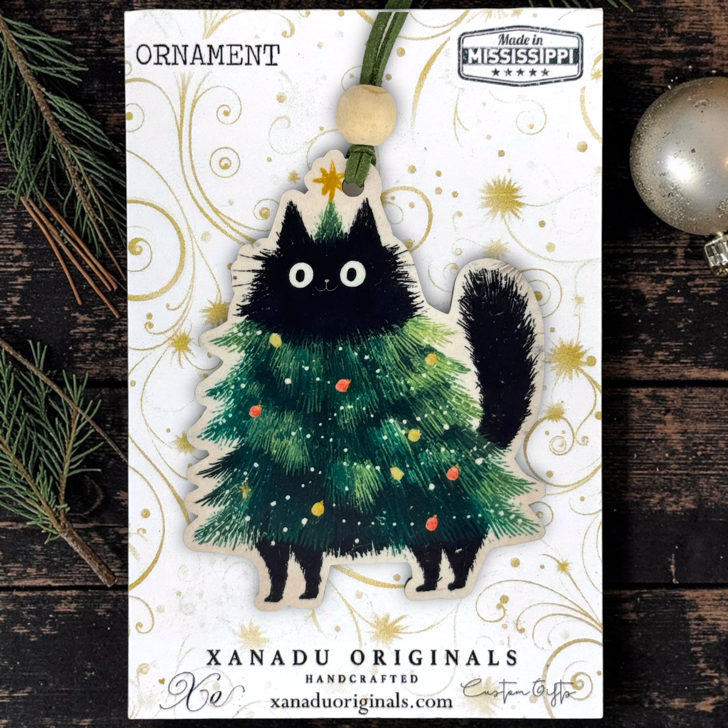 A Christmas ornament package from Xanadu Originals featuring a black cat dressed as a decorated Christmas tree. The ornament is displayed on a white card with gold swirls, with pine branches and a silver ornament on a dark wood background.