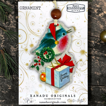 A Christmas ornament on a card featuring a handmade bird ornament with a red Santa hat, blue and pink feathers, a gold bell, a green wreath, red berries, and a light blue gift box with a red bow. The card is branded XANADU ORIGINALS and photographed on a dark rustic wood surface with pine needles.
