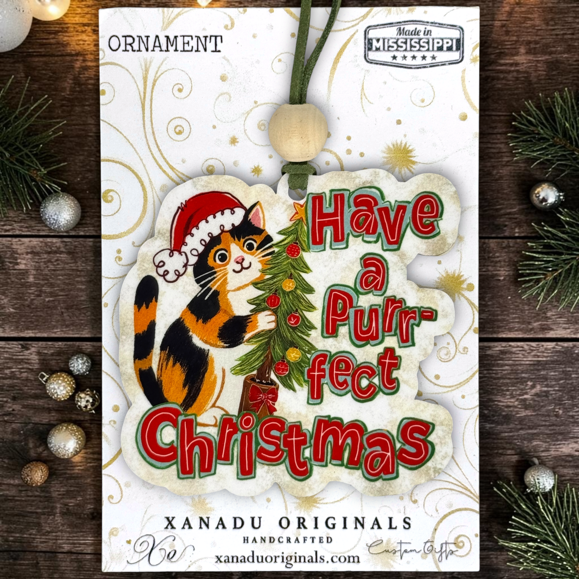 A Christmas ornament card featuring a cartoon calico cat wearing a Santa hat and licking a decorated Christmas tree, with the text “Have a Purrfect Christmas.” The card is photographed on a dark wood surface with pine branches and small ornaments around it.