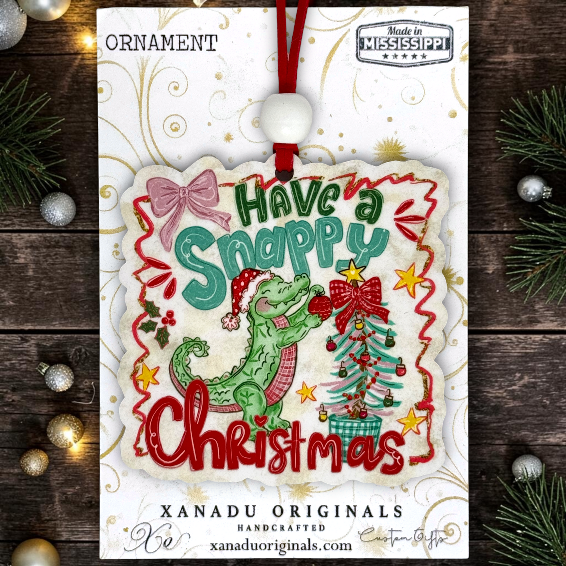 A Christmas ornament package from Xanadu Originals featuring a handcrafted ornament that reads 'Have a Snappy Christmas' with a green crocodile, red ribbon, pink bow, and a decorated Christmas tree. The ornament hangs from a red string on a white backing card, photographed on a dark wood surface with pine branches and ornaments around it.