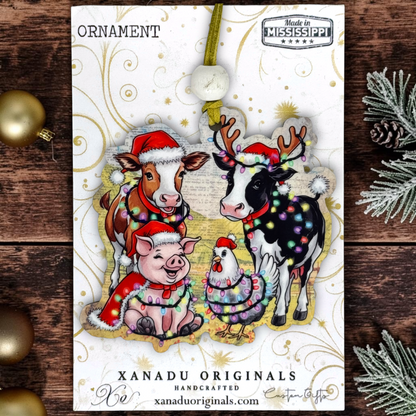 A Christmas ornament card featuring illustrated farm animals wearing Santa hats and holiday decorations, photographed on a rustic wood surface with gold ornaments and pine branches around it. The card shows the brand XANADU ORIGINALS and a 'Made in Mississippi' badge.
