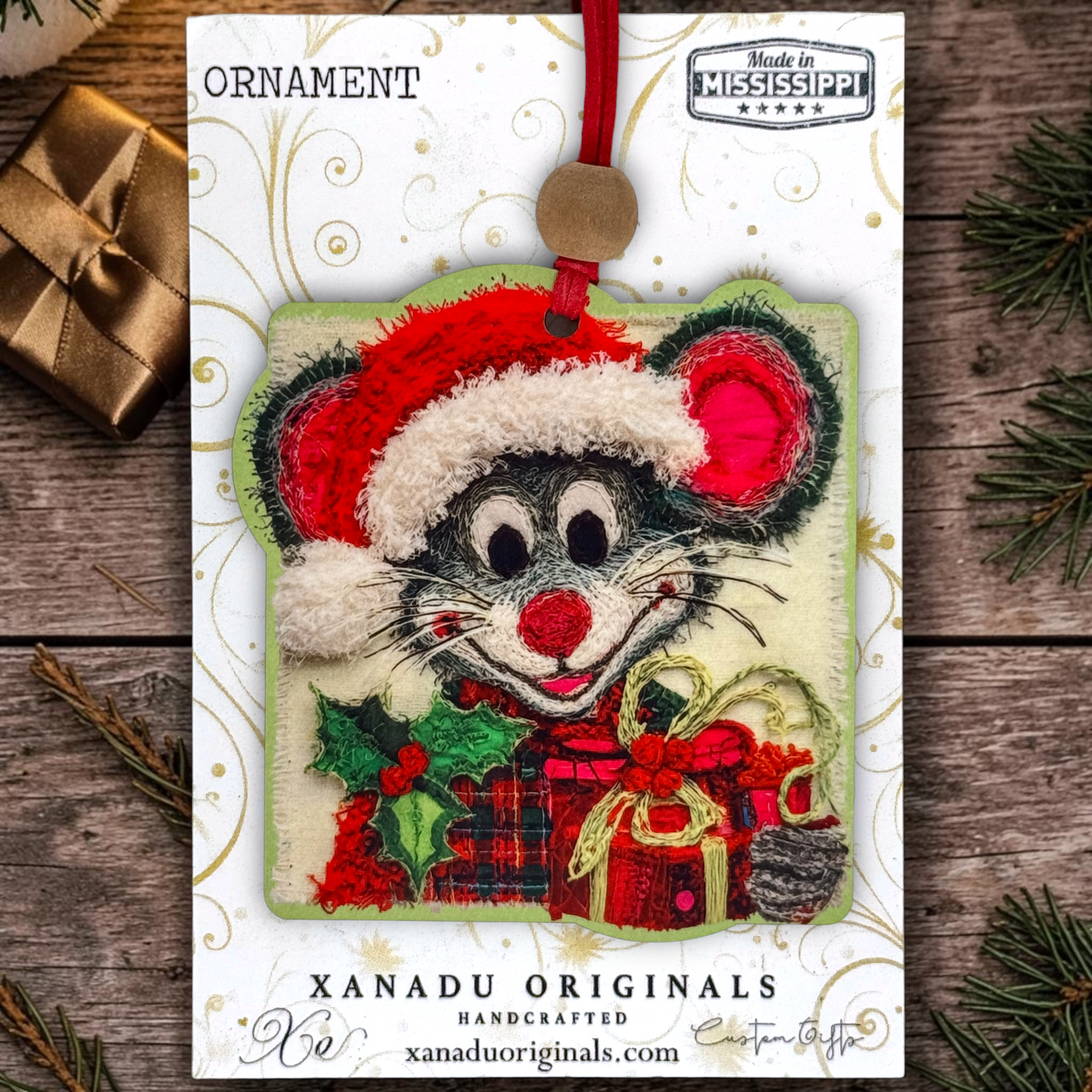 A handmade Christmas ornament featuring a gray mouse wearing a red Santa hat and holding wrapped gifts, displayed on its branded white backing card. The card reads 'ORNAMENT' and 'XANADU ORIGINALS HANDCRAFTED', with 'Made in Mississippi' at the top.