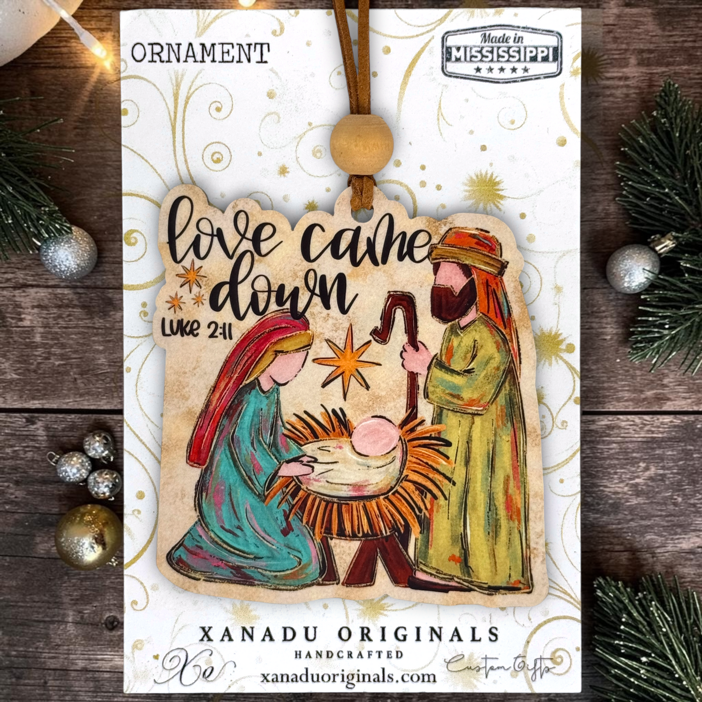 A Christmas ornament package from Xanadu Originals featuring a handcrafted wooden Nativity scene ornament. The ornament depicts Mary, Joseph, and baby Jesus in a manger with the text 'love came down Luke 2:11'.