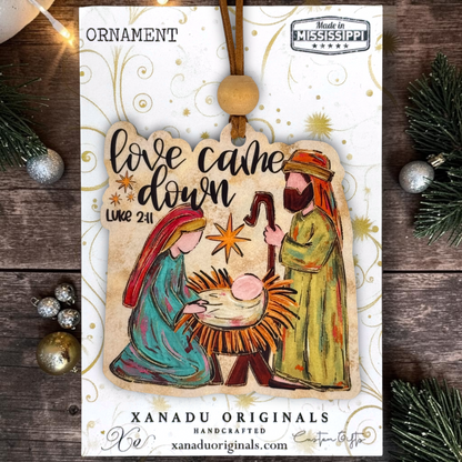 A Christmas ornament package from Xanadu Originals featuring a handcrafted wooden Nativity scene ornament. The ornament depicts Mary, Joseph, and baby Jesus in a manger with the text 'love came down Luke 2:11'.