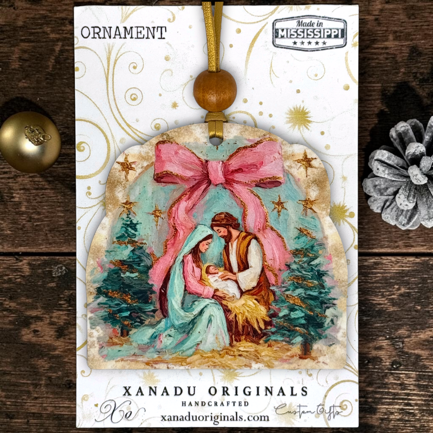 A Christmas ornament package from Xanadu Originals featuring a handcrafted ornament illustration of Mary, Joseph, and baby Jesus under a large pink bow, with gold decorative swirls and a 'Made in Mississippi' on a white background.