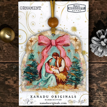 A Christmas ornament package from Xanadu Originals featuring a handcrafted ornament illustration of Mary, Joseph, and baby Jesus under a large pink bow, with gold decorative swirls and a 'Made in Mississippi' on a white background.