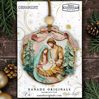 Handcrafted oval Christmas ornament featuring a nativity scene with Mary, Joseph, and baby Jesus, displayed on its white printed backing card with 'ORNAMENT' at the top and 'XANADU ORIGINALS' at the bottom. The card is styled on a dark wood surface with pine branches, pinecones, and gold ornaments around it.