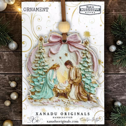 A handcrafted Christmas ornament by Xanadu Originals, featuring a nativity scene with Mary, Joseph, and baby Jesus under a pink bow, displayed on a rustic dark wood background with pine branches and silver ornaments.