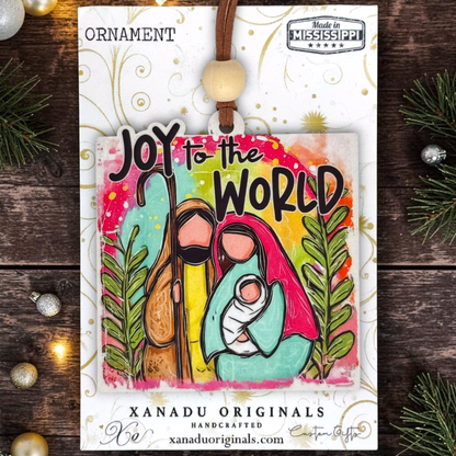 A square Christmas ornament on a card from Xanadu Originals featuring a colorful illustration of Mary, Joseph, and baby Jesus with the text 'JOY to the WORLD.' The ornament is displayed on a white backing card with a wooden bead loop and brown cord, set against a rustic dark wood background with pine branches and small ornaments.