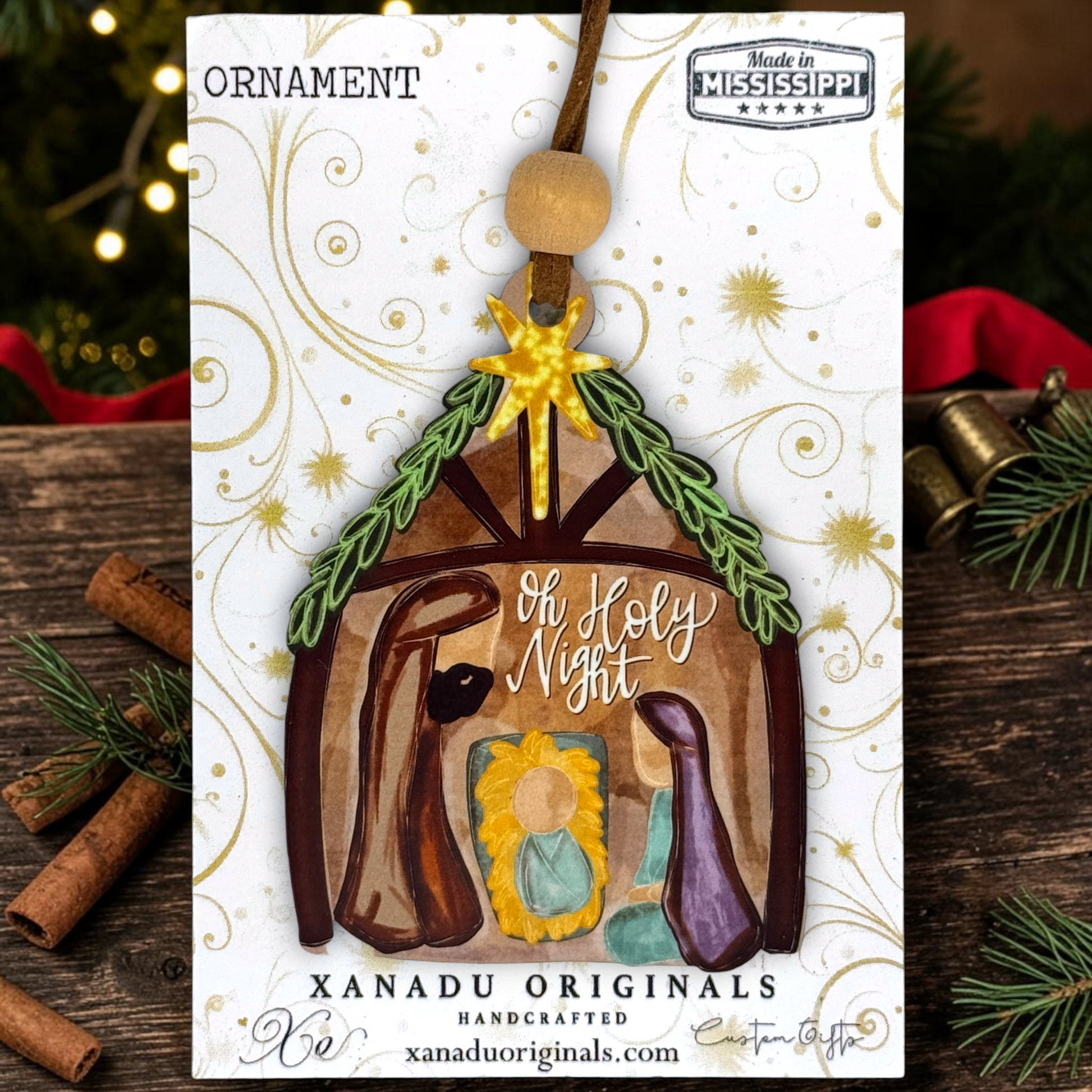 A handmade Christmas ornament shaped like a miniature stable with painted figures, displayed on its printed backing card. The card reads 'ORNAMENT' and 'XANADU ORIGINALS HANDCRAFTED', with 'Oh Holy Night' written on the ornament.