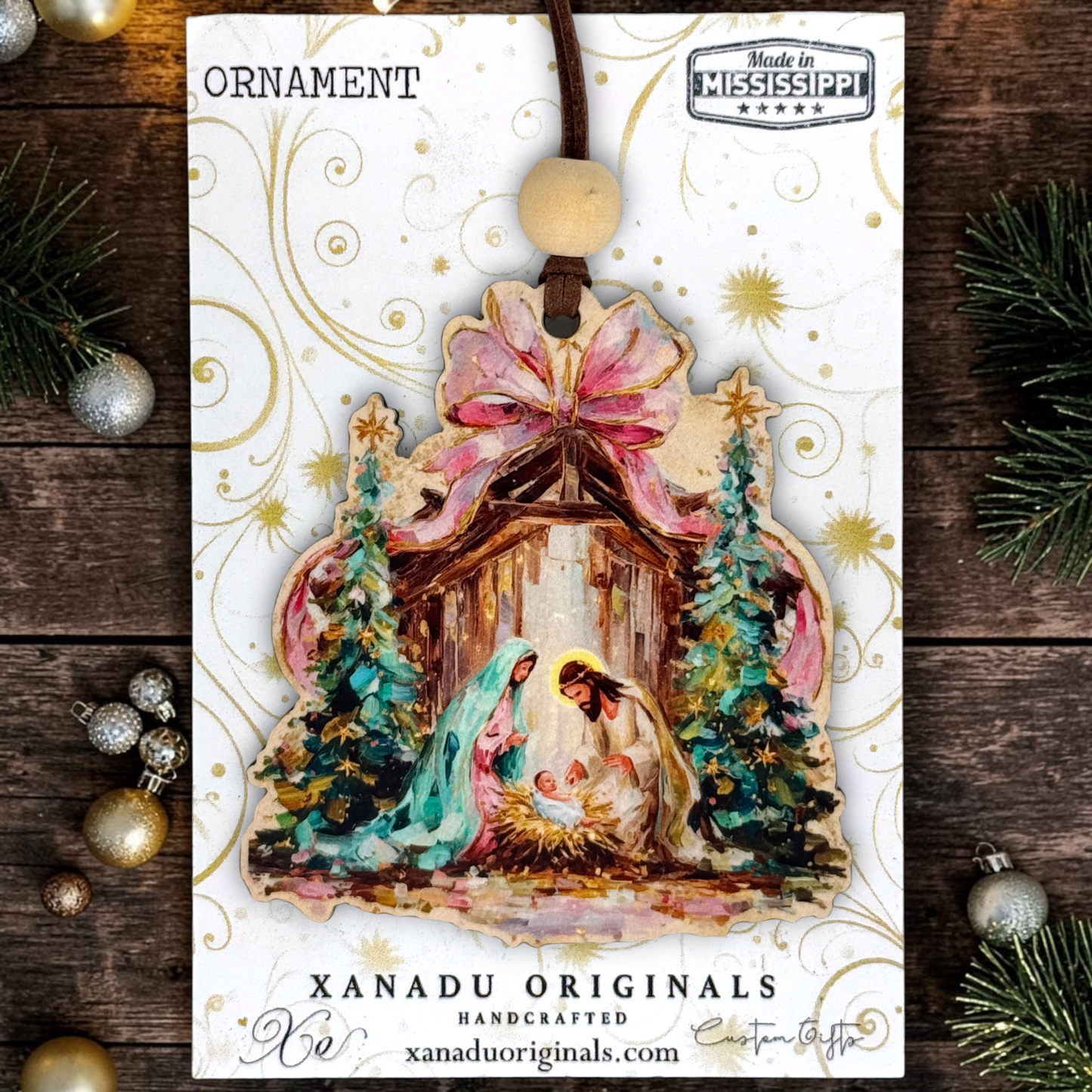 A Christmas ornament package from Xanadu Originals featuring a handcrafted nativity ornament with a pink bow, blue and green figures, and gold details on a white card, styled on a dark wood background with holiday ornaments and pine branches.