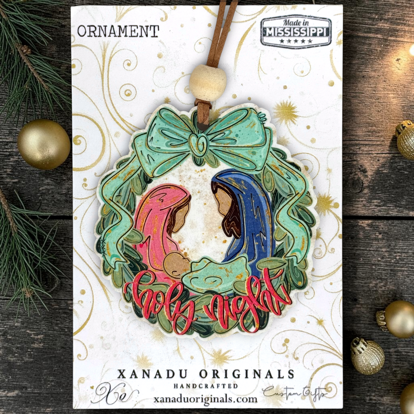 A Christmas ornament on a card featuring a handcrafted ornament design with Mary, Joseph, baby Jesus, a green wreath, and red 'Holy Night' lettering, shown on a branded card over a rustic wood background with pine branches and gold ornaments.