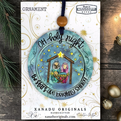 A handmade Christmas ornament card featuring a circular watercolor-style hanging ornament with a Nativity scene and the text “OH holy night the stars are brightly shining.” The card is branded XANADU ORIGINALS and includes a wooden bead ornament hanging from a dark cord, styled on a rustic wood surface with pine branches and gold ornaments.