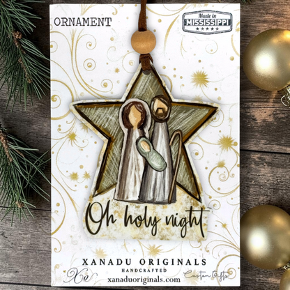 A handmade Christmas ornament card featuring a star-shaped nativity ornament with two figures and a baby, hanging from a wooden bead. The ornament reads 'Oh holy night' and shows 'Xanadu Originals' branding, styled on a rustic wood surface with pine branches and gold ornaments.
