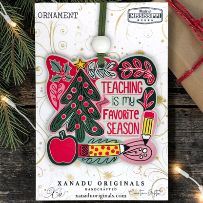 A Christmas ornament card featuring a colorful holiday design with a tree, apple, pencil, paintbrush, holly, and the phrase “TEACHING is my FAVORITE SEASON.” The card is branded XANADU ORIGINALS and shows “Made in Mississippi,” photographed on a rustic wood surface with string lights and paper packaging props around it.