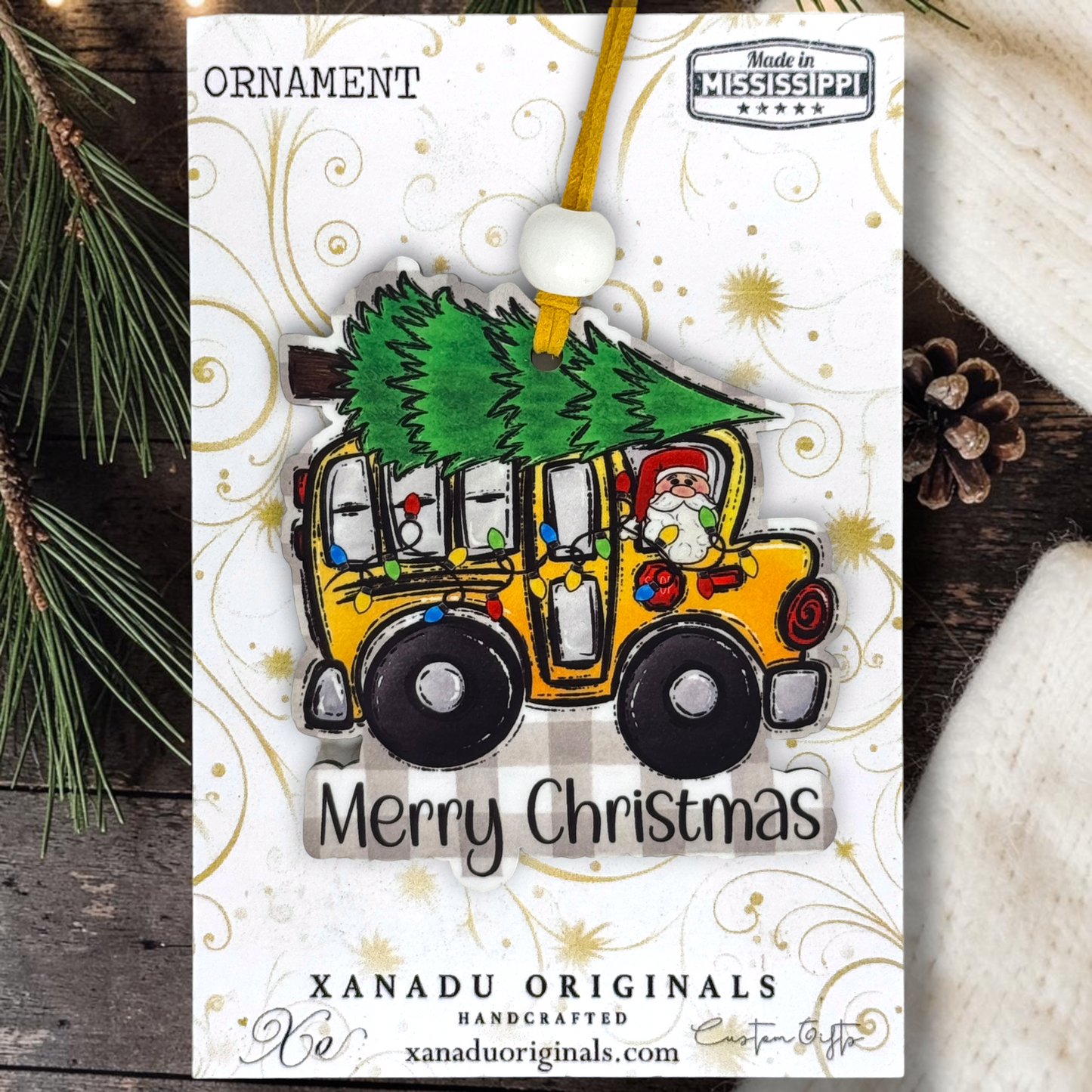 A Christmas ornament package from Xanadu Originals featuring a yellow jeep carrying a green Christmas tree, with a Santa figure in the driver's seat. The ornament is displayed on its white printed backing card with gold swirls, surrounded by pine branches and a pinecone.