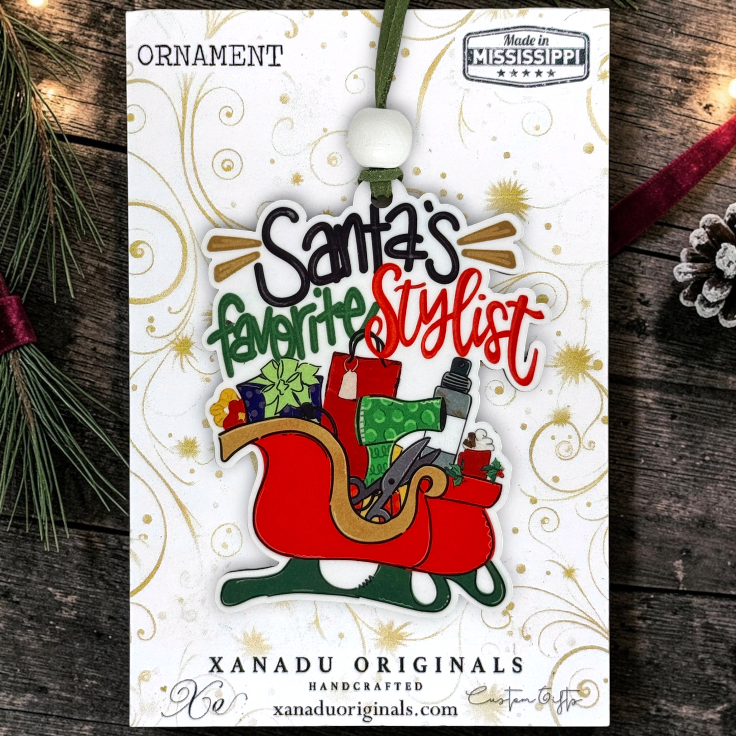 A Christmas ornament on its retail display card, featuring the text 'Santa's favorite Stylist' in black, green, and red lettering above an illustration of a red sleigh filled with gifts and a hat. The card is labeled 'ORNAMENT' and 'XANADU ORIGINALS HANDCRAFTED', and the product is photographed on a rustic dark wood surface with pine branches and a red ribbon.