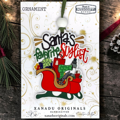 A Christmas ornament on its retail display card, featuring the text 'Santa's favorite Stylist' in black, green, and red lettering above an illustration of a red sleigh filled with gifts and a hat. The card is labeled 'ORNAMENT' and 'XANADU ORIGINALS HANDCRAFTED', and the product is photographed on a rustic dark wood surface with pine branches and a red ribbon.