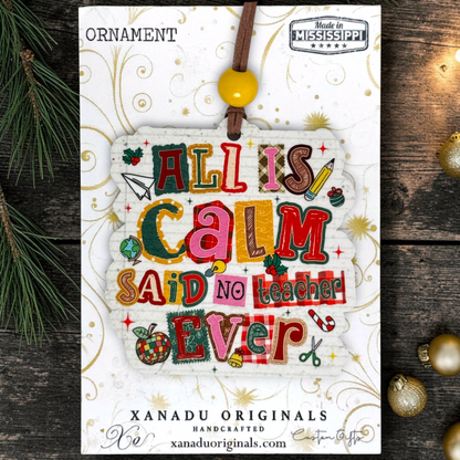 A handmade Christmas ornament card featuring the phrase 'ALL IS CALM SAID NO TEACHER EVER' in colorful patchwork letters, hanging from a brown cord with a yellow bead. The card is branded 'XANADU ORIGINALS' and shows 'Made in Mississippi' at the top, styled on a dark wood background with pine branches and gold ornaments.