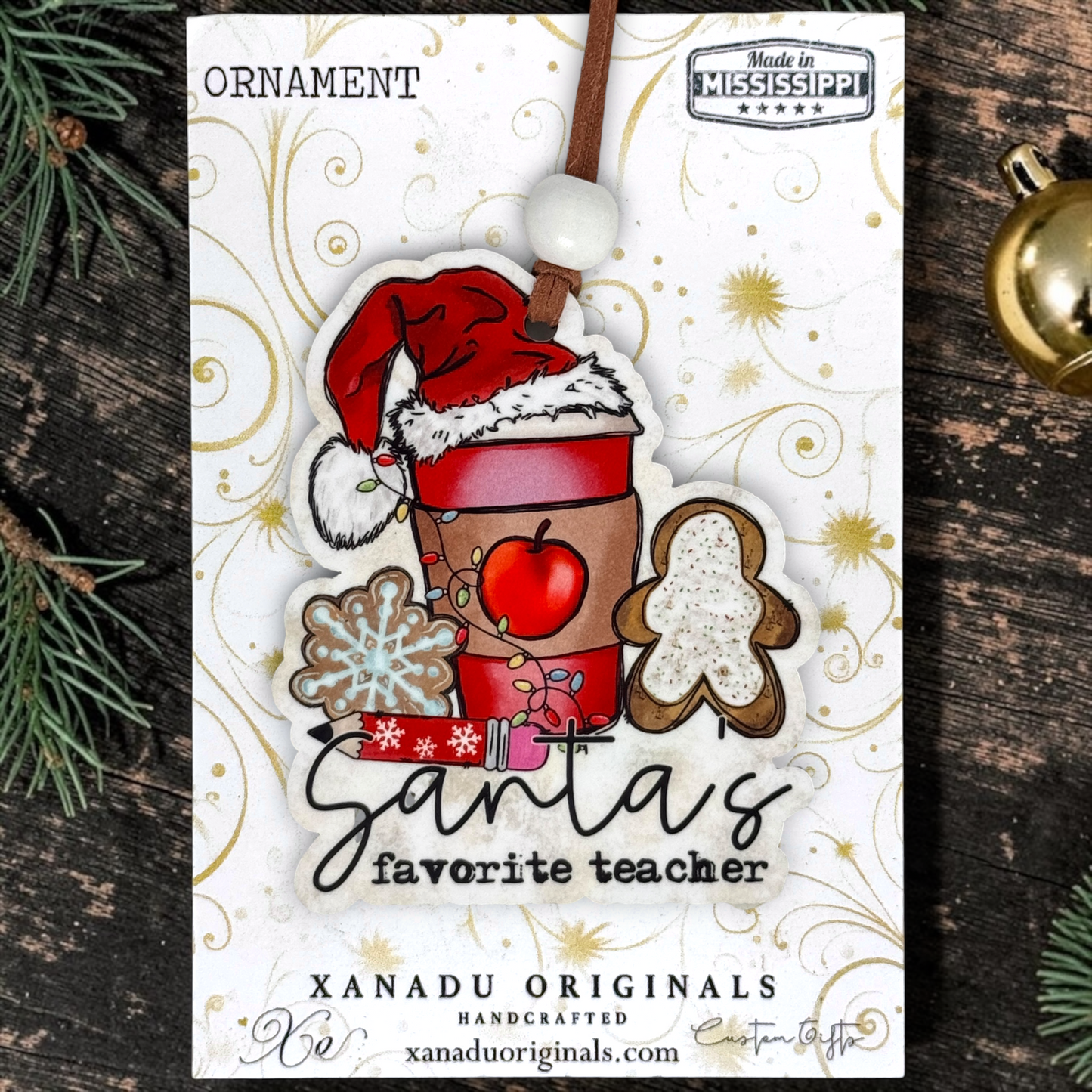 A Christmas ornament on a card featuring a festive ornament design with the text 'Santa's favorite teacher,' surrounded by holiday motifs like a Santa hat, gingerbread figure, snowflake, apple, and gift. The card is photographed on a dark wood surface with pine branches and a gold ornament nearby.