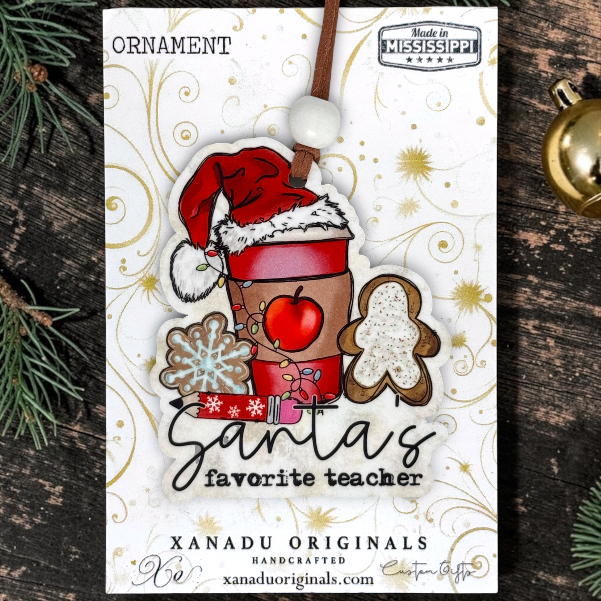A Christmas ornament on a card featuring a festive ornament design with the text 'Santa's favorite teacher,' surrounded by holiday motifs like a Santa hat, gingerbread figure, snowflake, apple, and gift. The card is photographed on a dark wood surface with pine branches and a gold ornament nearby.