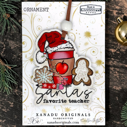 A Christmas ornament on a card featuring a festive ornament design with the text 'Santa's favorite teacher,' surrounded by holiday motifs like a Santa hat, gingerbread figure, snowflake, apple, and gift. The card is photographed on a dark wood surface with pine branches and a gold ornament nearby.