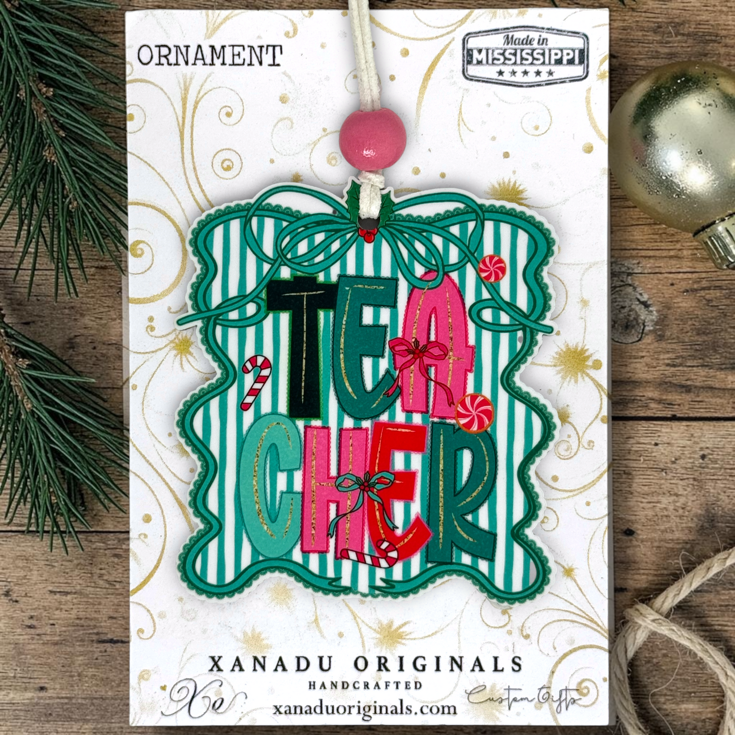 A festive Christmas ornament card featuring the word 'TEACHER' in colorful letters with candy cane and ribbon accents, hanging from a pink bead on a printed card. The card is photographed on a rustic wood surface with pine branches and a silver ornament visible at the edges.