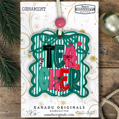 A festive Christmas ornament card featuring the word 'TEACHER' in colorful letters with candy cane and ribbon accents, hanging from a pink bead on a printed card. The card is photographed on a rustic wood surface with pine branches and a silver ornament visible at the edges.