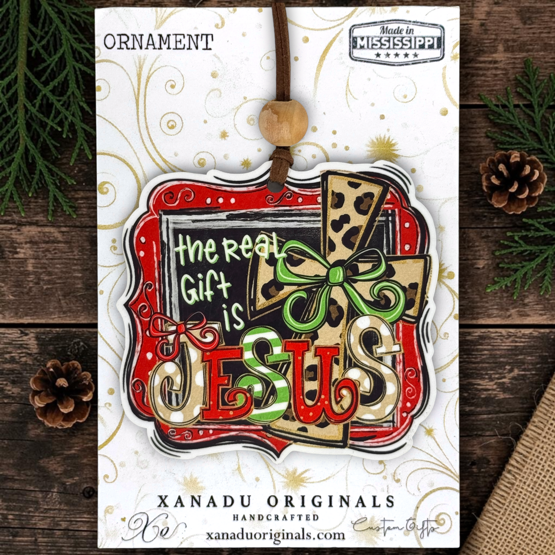 A Christmas ornament on a card featuring a layered ornament design with the words “The Real Gift is JESUS,” a leopard-print cross, and a green bow. The card is branded XANADU ORIGINALS and shows the product on a rustic wood background with pine branches and pinecones.