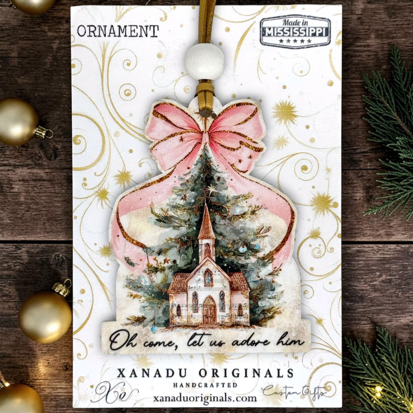 A Christmas ornament card featuring a handcrafted ornament design with a pink bow, gold glitter accents, a church illustration, and the text 'Oh, come, let us adore him.' The card is branded XANADU ORIGINALS and includes a 'Made in Mississippi' badge, shown on a rustic wood background with holiday decorations.
