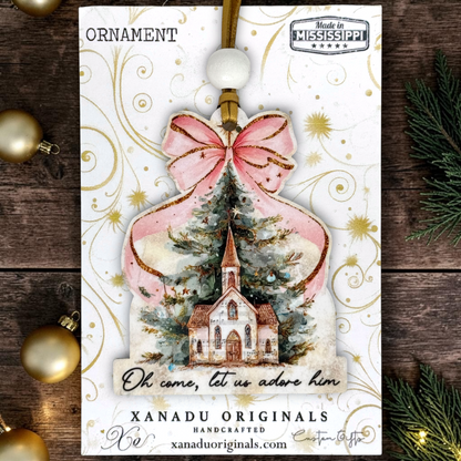 A Christmas ornament card featuring a handcrafted ornament design with a pink bow, gold glitter accents, a church illustration, and the text 'Oh, come, let us adore him.' The card is branded XANADU ORIGINALS and includes a 'Made in Mississippi' badge, shown on a rustic wood background with holiday decorations.