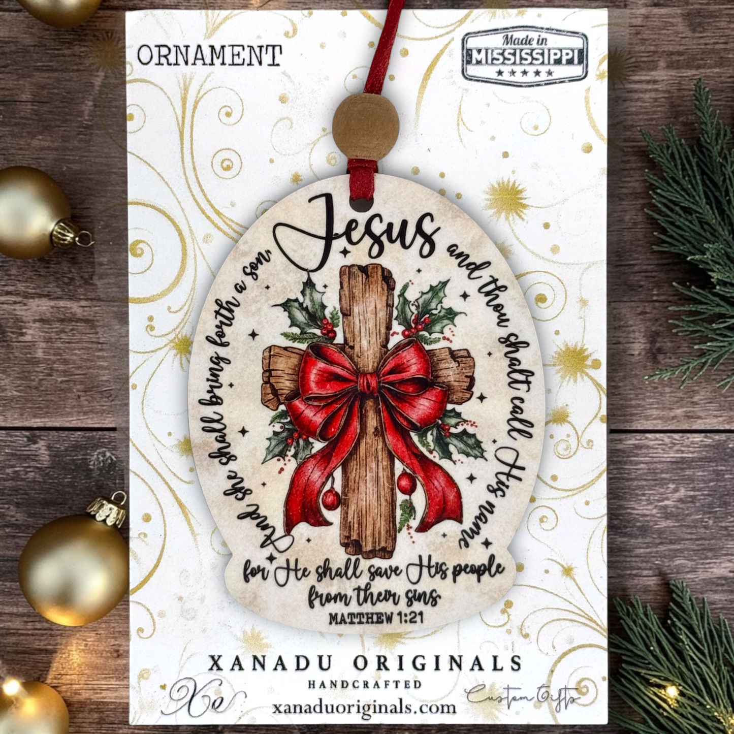 A Christmas ornament card featuring a round ornament design with a wooden cross, red bow, holly leaves, and scripture text. The card reads 'ORNAMENT' at the top and shows 'XANADU ORIGINALS' branding, with gold decorative swirls on a white background.