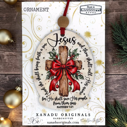 A Christmas ornament card featuring a round ornament design with a wooden cross, red bow, holly leaves, and scripture text. The card reads 'ORNAMENT' at the top and shows 'XANADU ORIGINALS' branding, with gold decorative swirls on a white background.