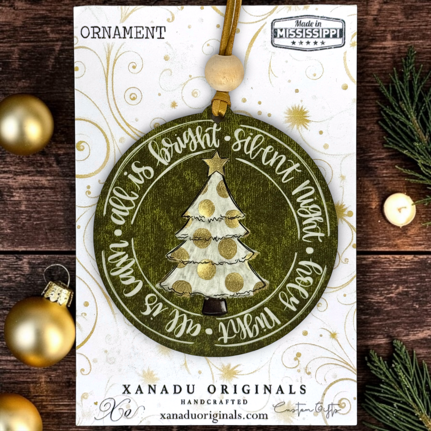 A round Christmas ornament card featuring a handcrafted olive green ornament with a white and gold glittery tree, surrounded by the phrase 'All is bright, silent night, peace with God.' The card is branded XANADU ORIGINALS and shows a wooden bead hanging loop.