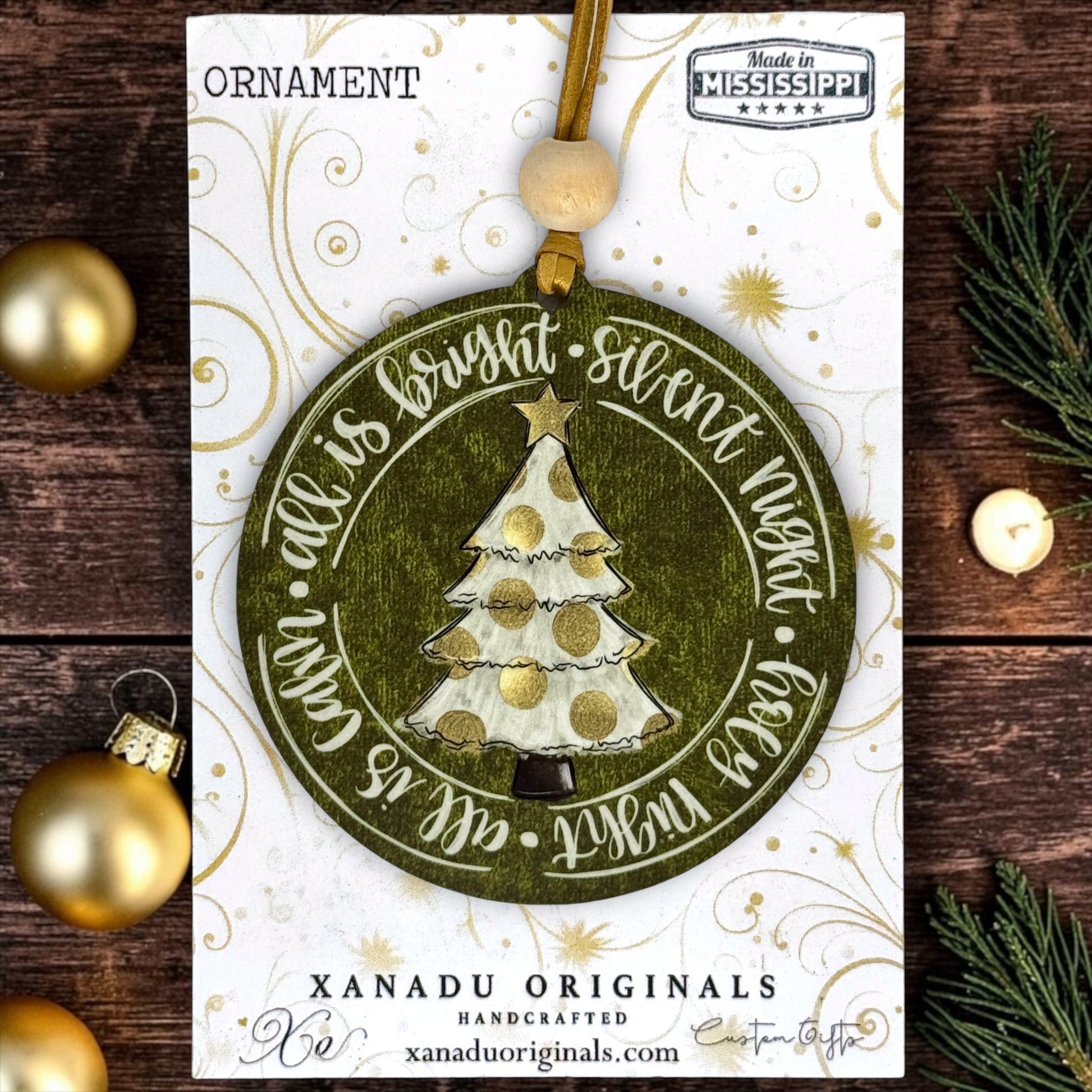 A round Christmas ornament card featuring a handcrafted olive green ornament with a white and gold glittery tree, surrounded by the phrase 'All is bright, silent night, peace with God.' The card is branded XANADU ORIGINALS and shows a wooden bead hanging loop.