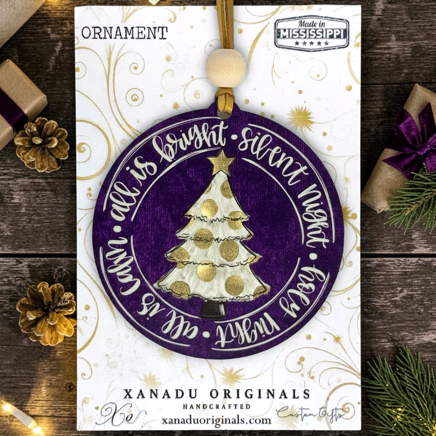 A circular hanging ornament displayed on its printed backing card. The ornament is purple with a white and gold Christmas tree in the center and white script text around the edge.