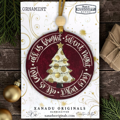 A round Christmas ornament carded on a decorative display card. The ornament is dark red with a hand-painted white and gold tree in the center and white script text around the edge.