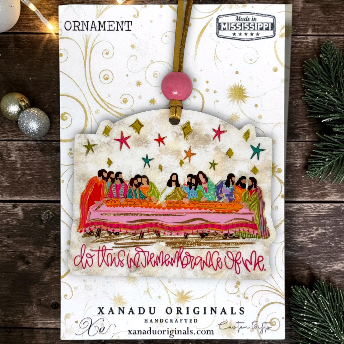 A Christmas ornament card from Xanadu Originals featuring a handcrafted ornament illustration with a colorful depiction of the Last Supper, gold swirls, and a pink hanging bead, displayed on a rustic wooden background with holiday decorations.