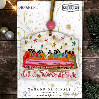 A Christmas ornament card from Xanadu Originals featuring a handcrafted ornament illustration with a colorful depiction of the Last Supper, gold swirls, and a pink hanging bead, displayed on a rustic wooden background with holiday decorations.