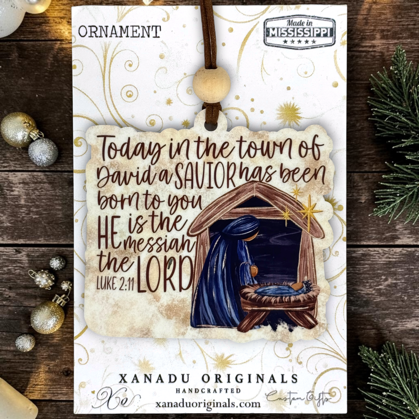 A Christmas ornament on a card from Xanadu Originals featuring a hanging ornament design with a nativity scene and the text, 'Today in the town of David a SAVIOR has been born to you HE is the messiah the LORD Luke 2:11.' The card is styled on a rustic dark wood surface with holiday ornaments and evergreen branches around it.