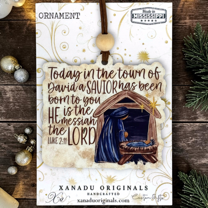 A Christmas ornament on a card from Xanadu Originals featuring a hanging ornament design with a nativity scene and the text, 'Today in the town of David a SAVIOR has been born to you HE is the messiah the LORD Luke 2:11.' The card is styled on a rustic dark wood surface with holiday ornaments and evergreen branches around it.