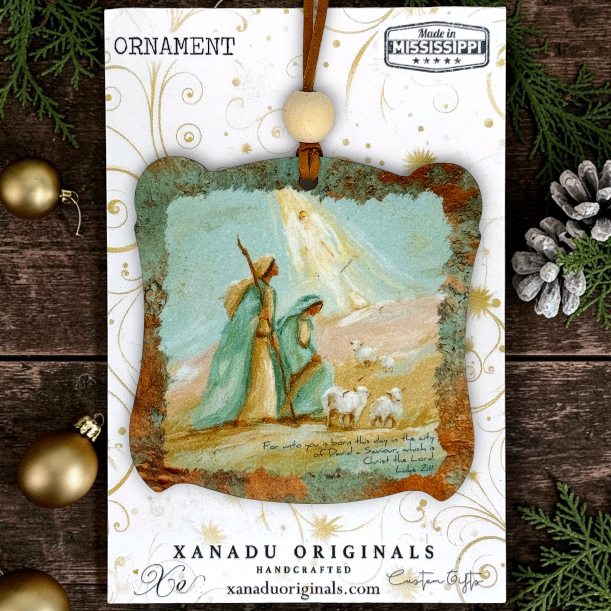 A Christmas ornament card featuring a handcrafted, rustic wood ornament with a painted wise men and angel with sheep. The card is branded 'XANADU ORIGINALS' and decorated with gold swirls on a white background, surrounded by pine branches, a pinecone, and gold ornaments on a dark wood surface.