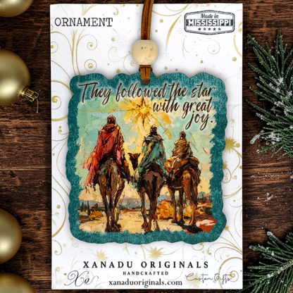 A Christmas ornament on a card featuring a handcrafted ornament with an illustration of three camels and wise men under a glowing star, with the text 'They followed the star with great joy.' The card is branded XANADU ORIGINALS and photographed on a rustic wooden surface with gold ornaments and evergreen branches around it.