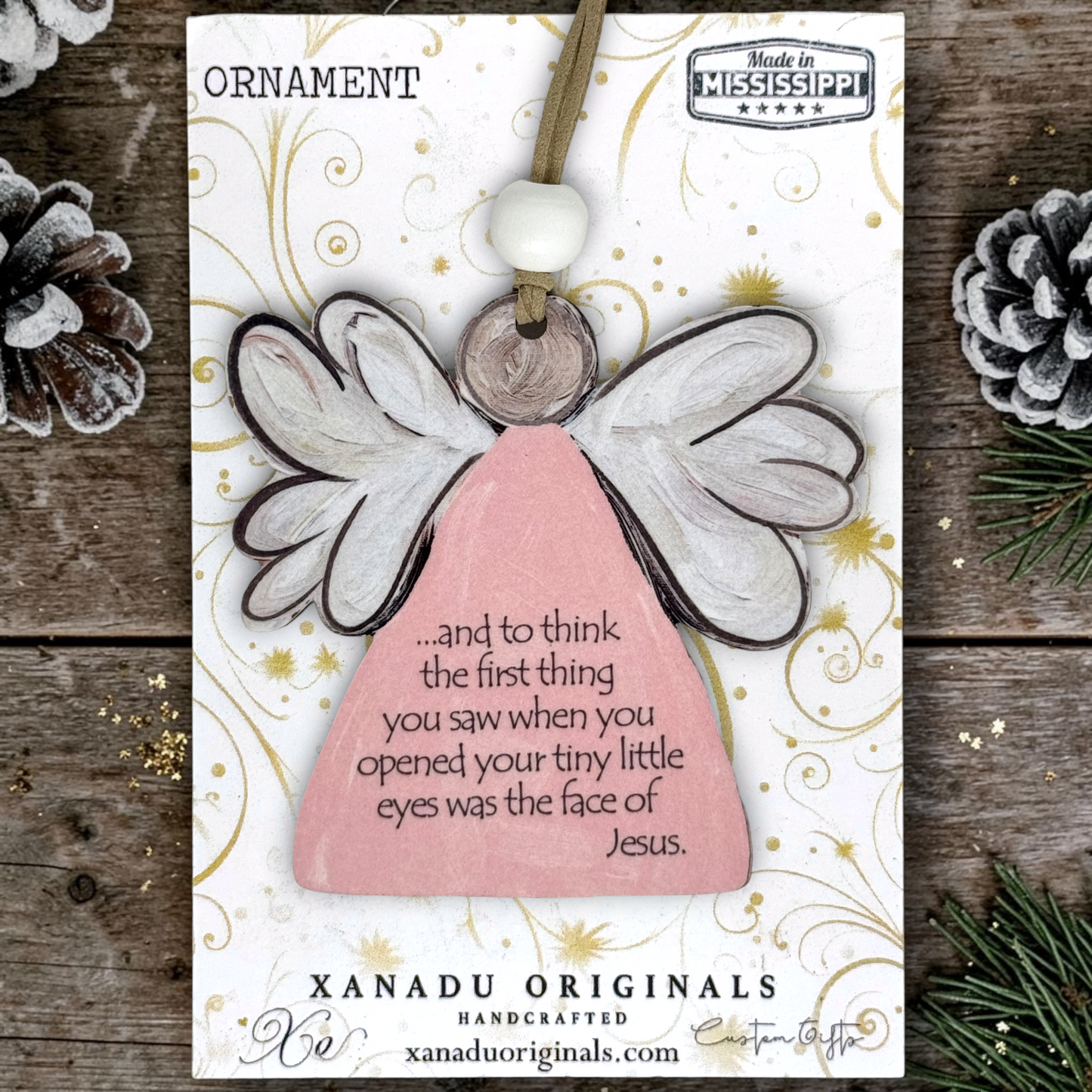 A wooden angel ornament on a printed display card, photographed flat on a rustic wood surface with pinecones and evergreen branches around it. The ornament has a pale pink body, white wings with dark outlines, a white bead, and a small wooden disc.